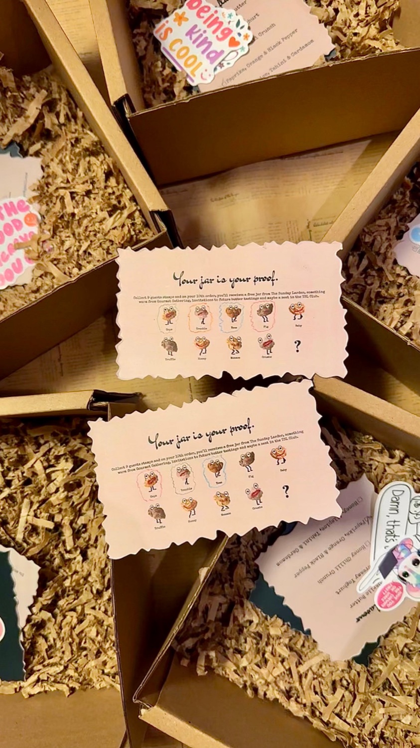 Pantry Passport stamp cards resting on open Sunday Larder boxes packed with shredded kraft paper and stickers